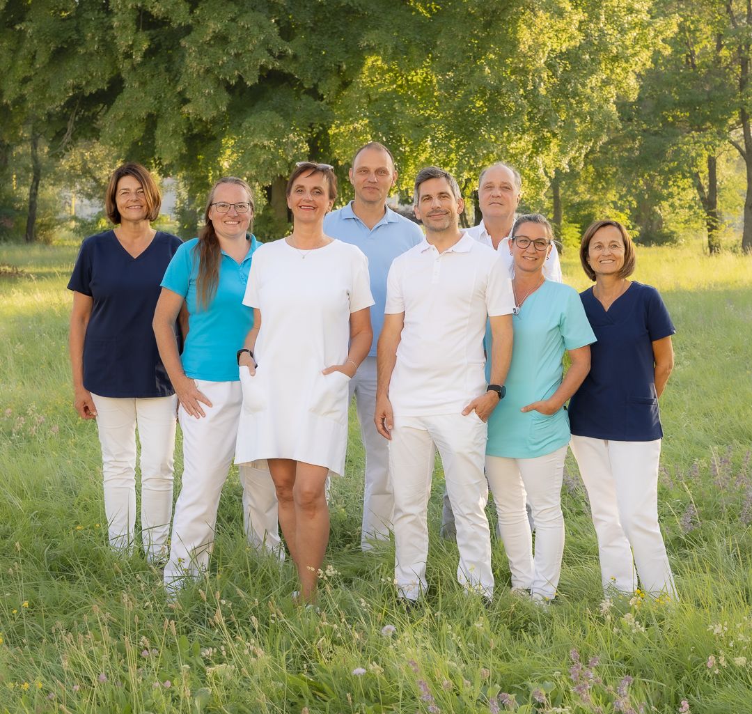 Das Ortho-Vital-Team – Ärztin, Ärzte, Therapeuten und Assistentinnen – steht gemeinsam auf einer sommerlichen Wiese vor Bäumen